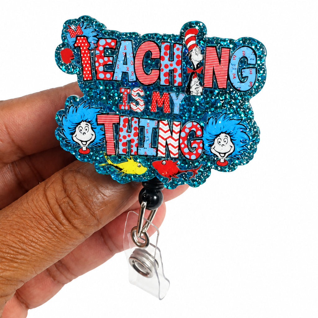 A hand holding a glittery cloud-shaped badge reel with the text 'TEACHING IS MY THING' in pink and blue lettering, featuring cartoon characters and a striped hat on a white background.