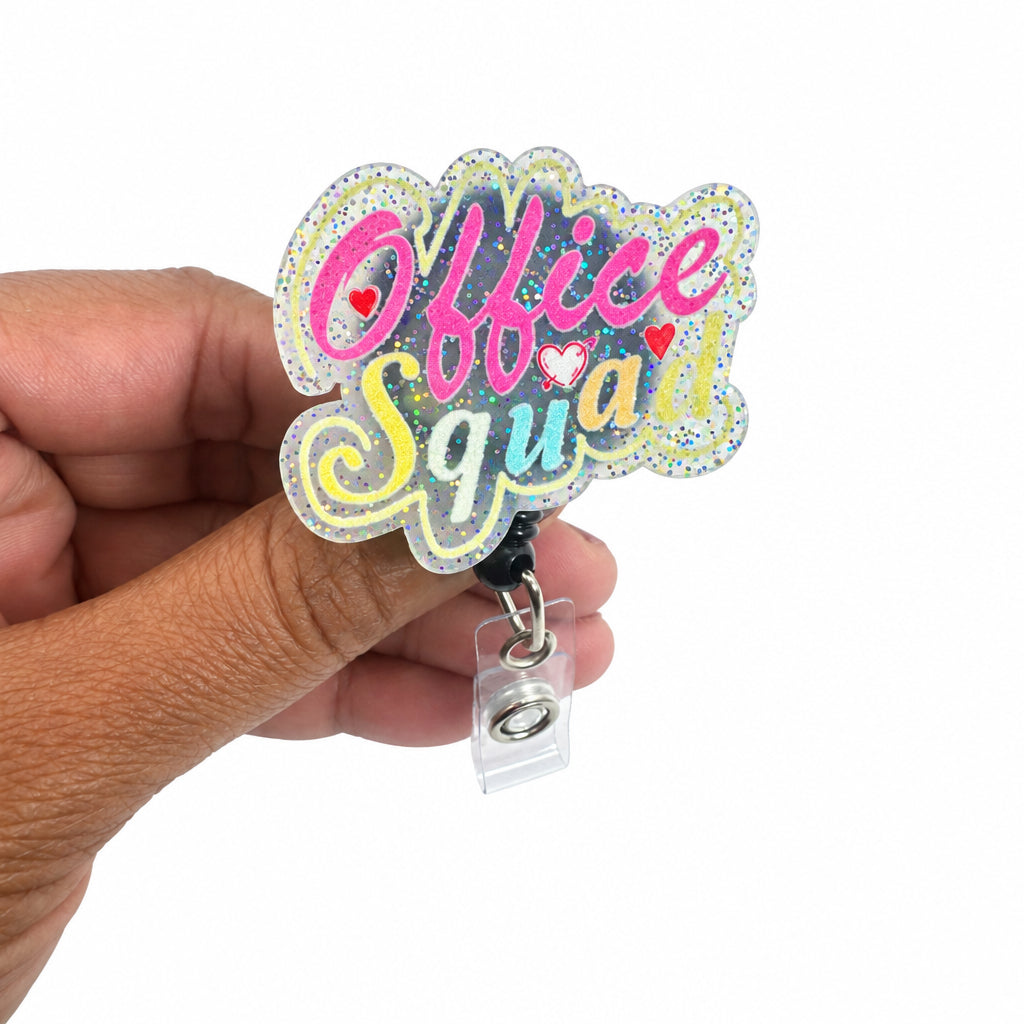 A hand holding a die-cut acrylic badge reel sign that reads 'Office Squad' in pink, yellow, and white glittery lettering, with a clear plastic clip at the bottom, against a plain white background.