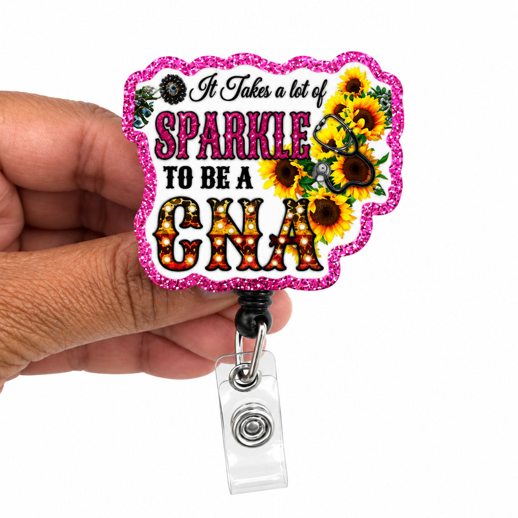 A hand holding a pink glitter badge reel with a decorative patch that reads 'It Takes a lot of SPARKLE TO BE A GNA' with sunflower graphics, on a plain white background.