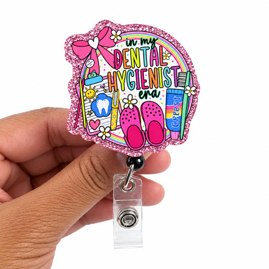 A hand holding a die-cut acrylic badge reel cover featuring a colorful cartoon illustration of dental hygiene supplies and the text 'In my DENTAL HYGIENIST JOB!'. The badge reel is pink glitter-edged with a clear plastic clip attached.
