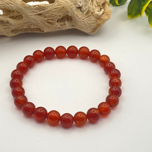 Round red beaded bracelet on a white surface with natural wood and green leaves in the background