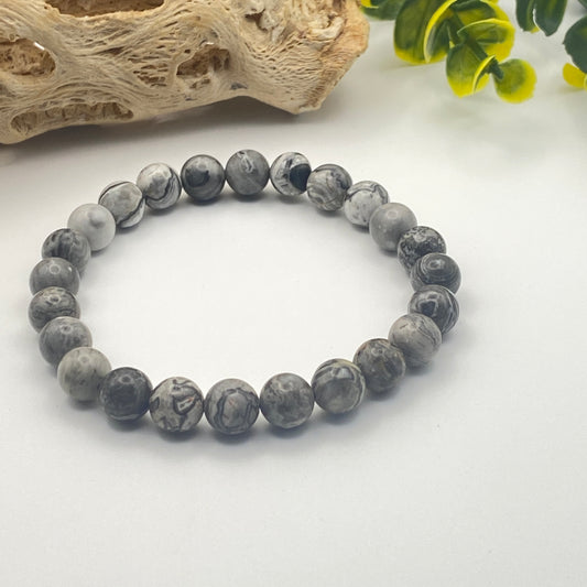 Gray beaded bracelet on a white surface with natural wood and green leaves in the background