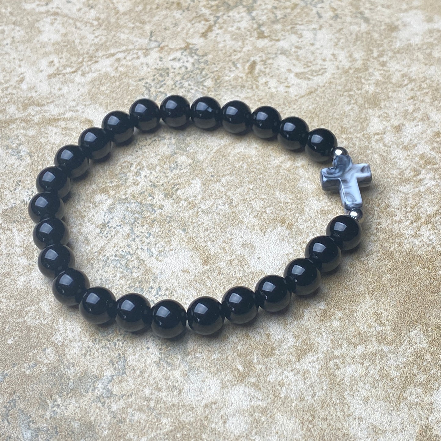 Black beaded bracelet with a silver cross charm on a textured surface