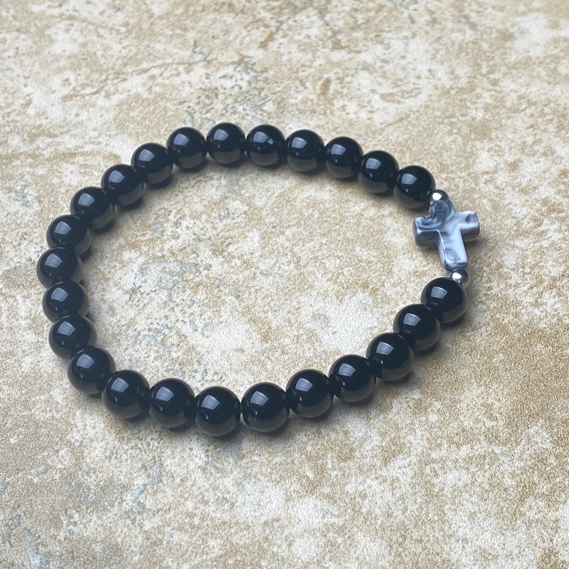 Black beaded bracelet with a silver cross charm on a textured surface