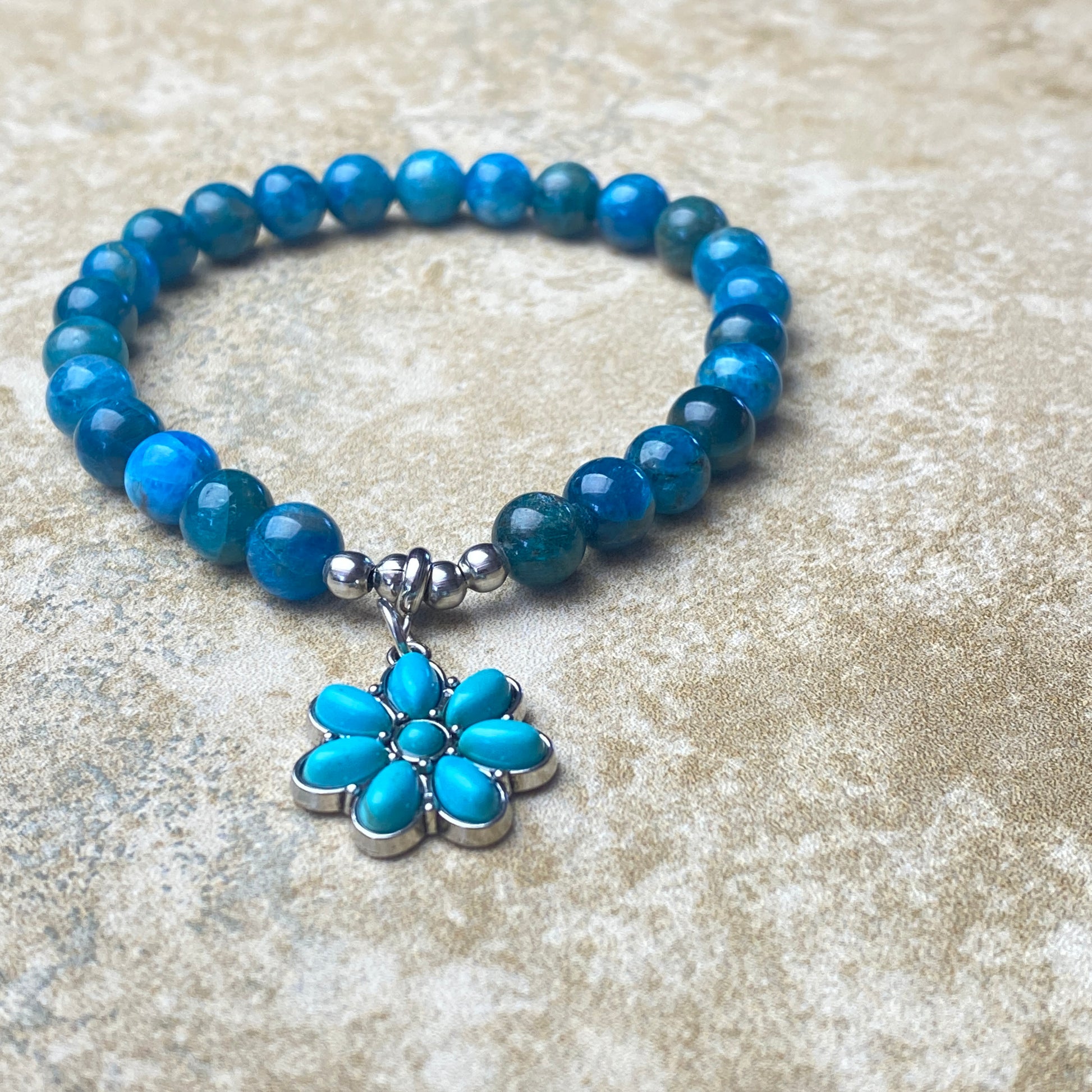 Blue beaded bracelet with a silver and blue flower charm on a textured surface