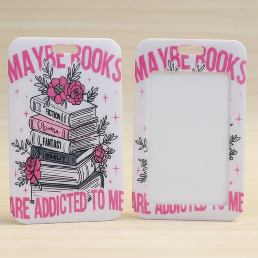 Two views of a white phone case with a pink and black book-themed design, standing on a light wood surface. The left case shows a stack of books with pink roses and the text 'MAYBE BOOKS ARE ADDICTED TO ME', while the right case shows a blank white back.