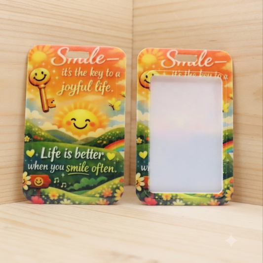 Two small rectangular photo frames with rounded corners displayed side by side on a light wood surface against a wood-paneled background. Each frame has a colorful printed border featuring a smiling sun, flowers, grass, and the phrase 'Smile it's the key to a joyful life.