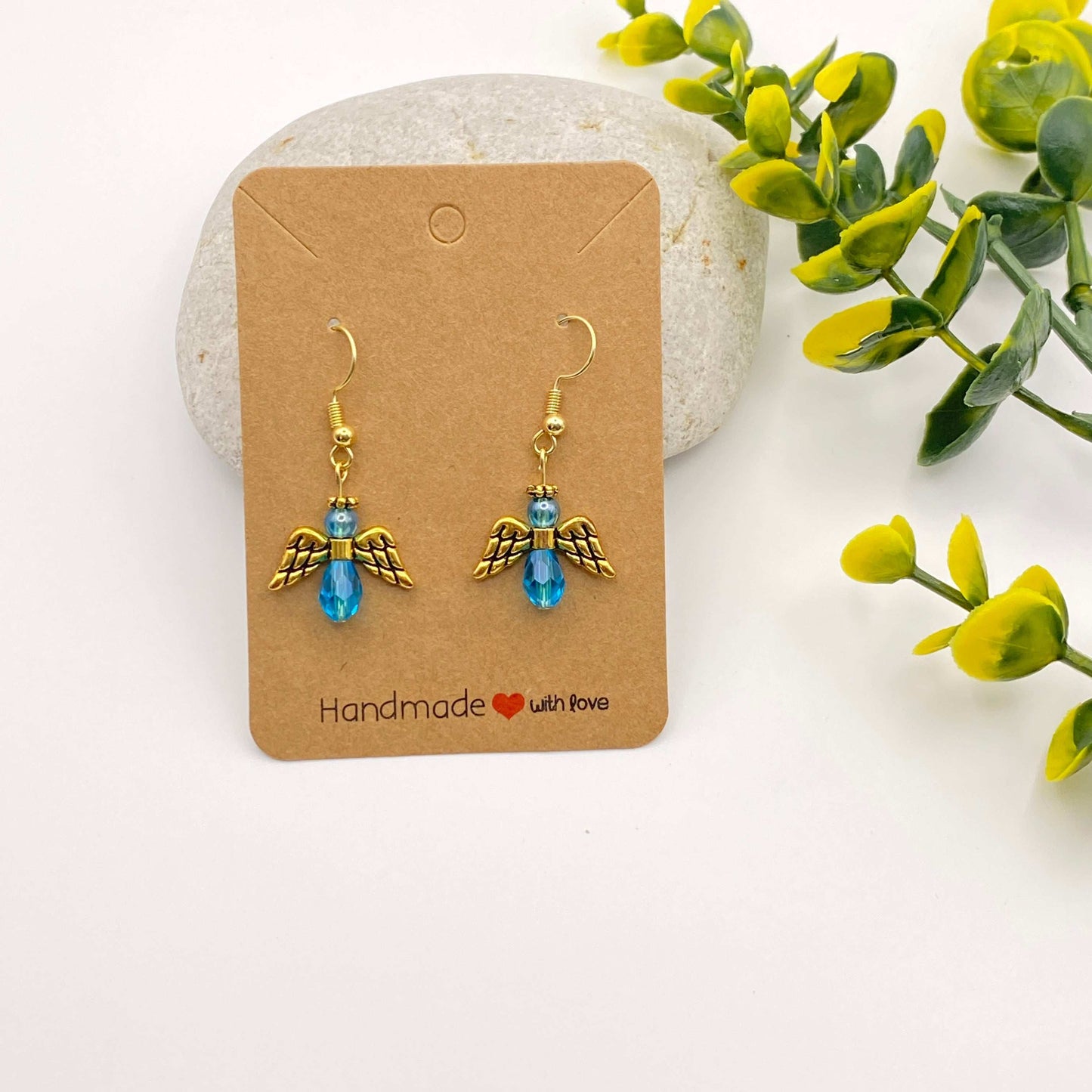 Gold angel earrings with blue stones on a 'Handmade with Love' card, surrounded by greenery.