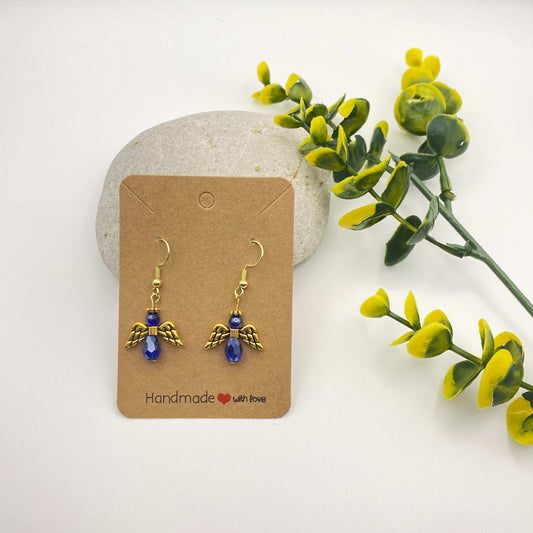 Angel-shaped earrings on a cardboard backing with a natural stone and greenery in the background