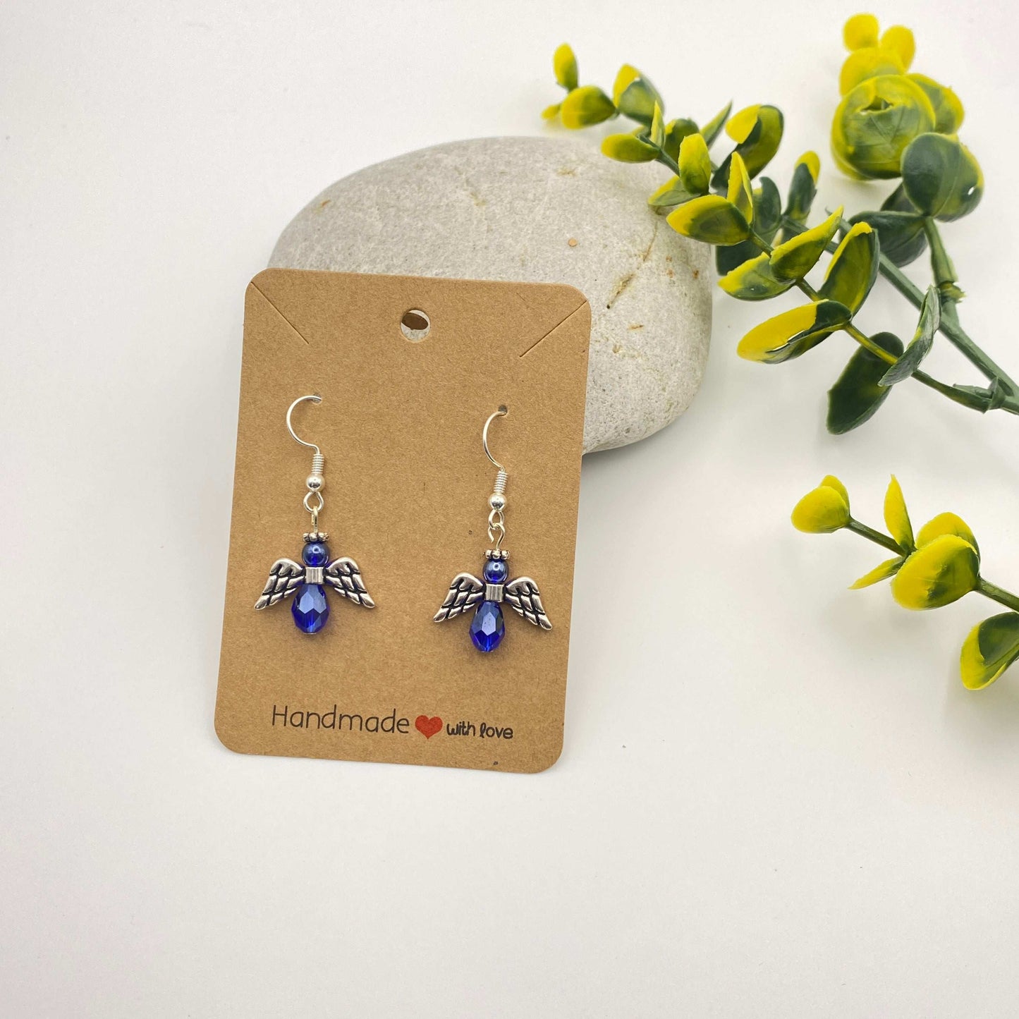 Pair of angel-shaped earrings with blue gemstones on a 'Handmade with Love' card, surrounded by greenery.