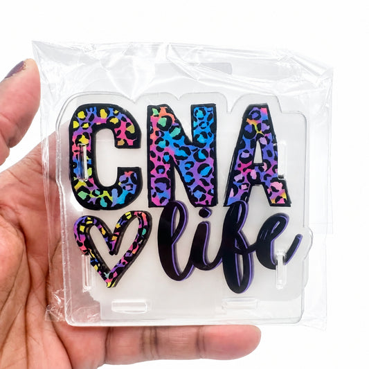 A hand holding a clear acrylic plaque that reads 'GNA life' in a colorful leopard print and black script font, still inside its clear plastic packaging.