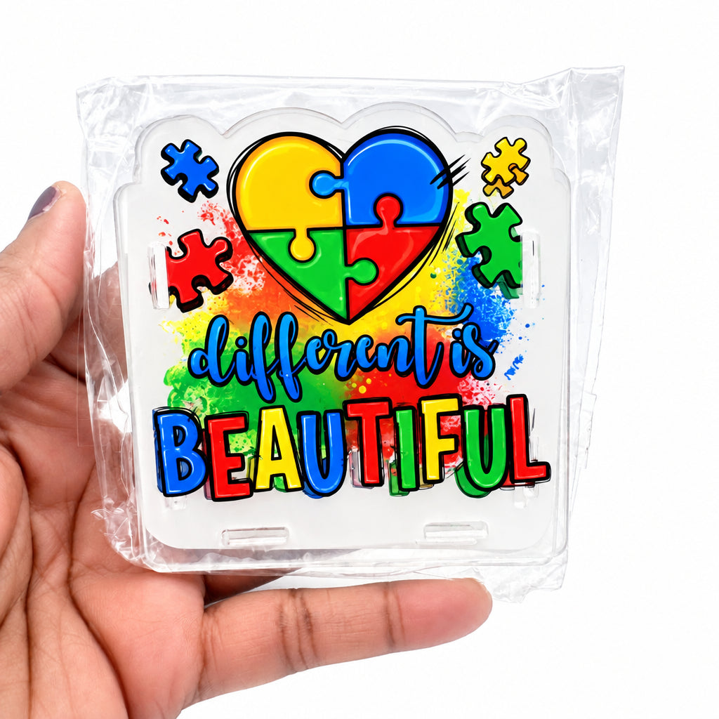 A hand holding a clear plastic-wrapped square sticker or decal featuring a puzzle-heart design and the words 'different is BEAUTIFUL' in colorful lettering on a white background.