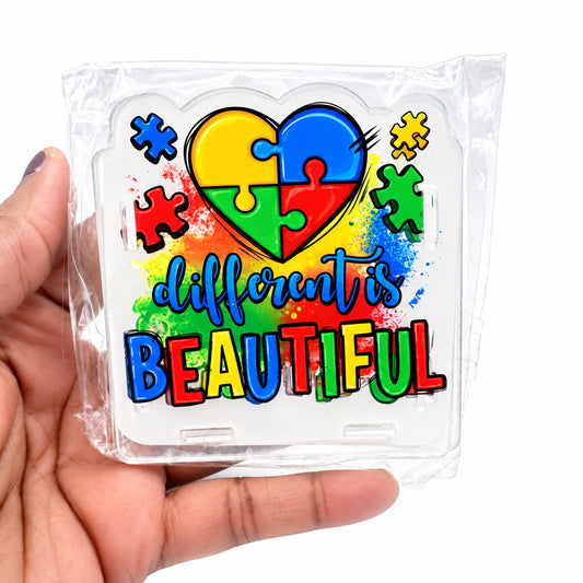 A hand holding a clear plastic-wrapped square sticker or decal featuring a puzzle-heart design and the words 'different is BEAUTIFUL' in colorful lettering on a white background.