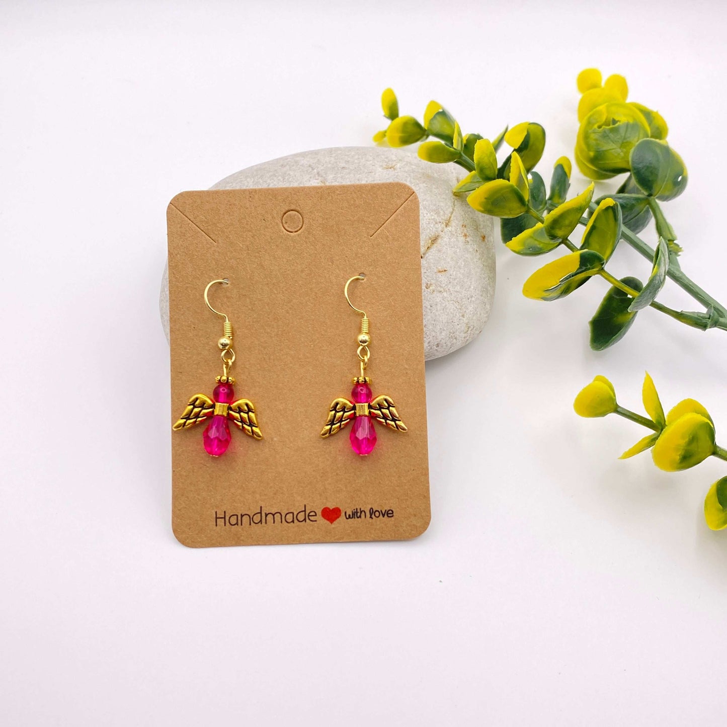 Pair of gold angel wing earrings with pink beads on a 'Handmade with Love' card, surrounded by greenery.