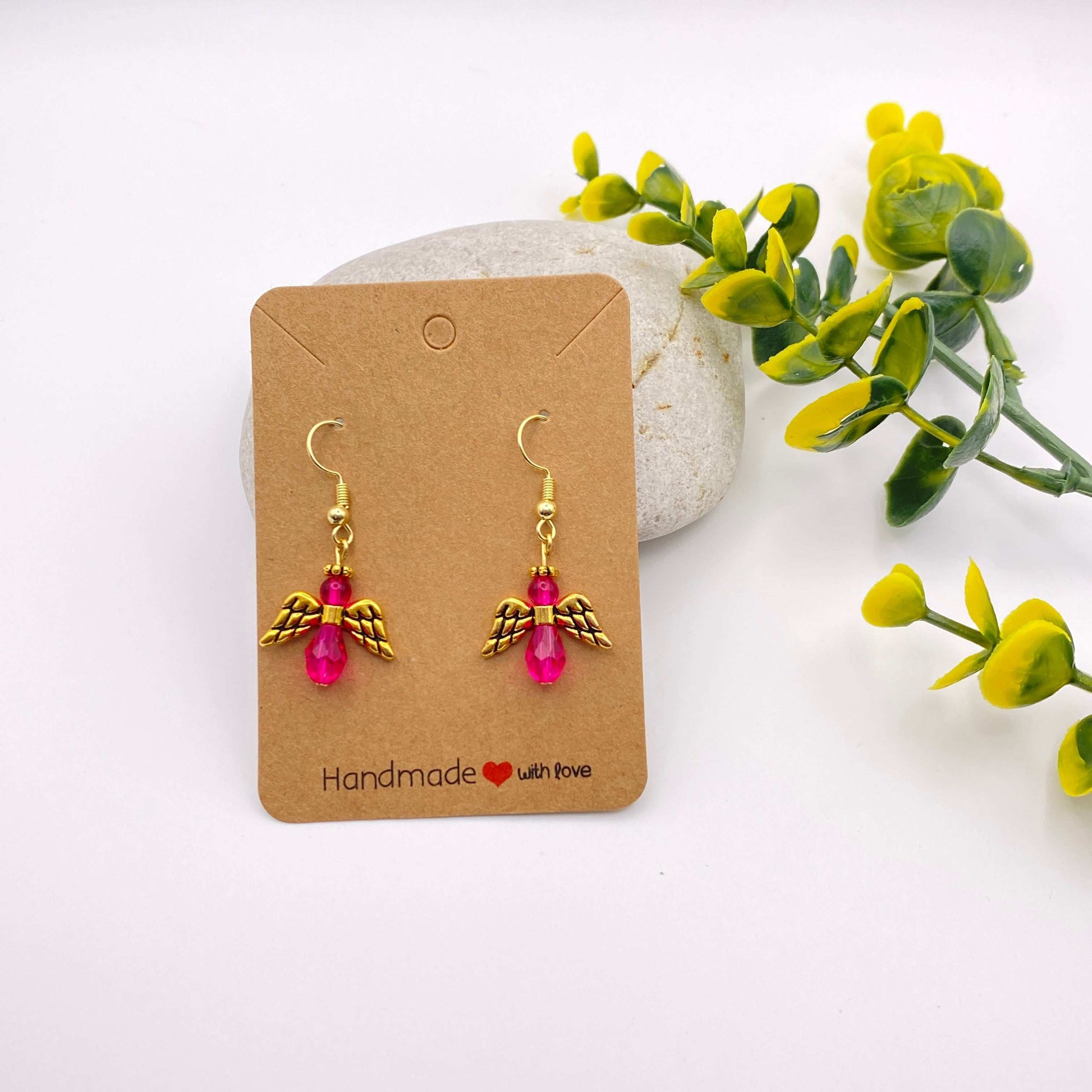 Pair of gold angel wing earrings with pink beads on a 'Handmade with Love' card, surrounded by greenery.