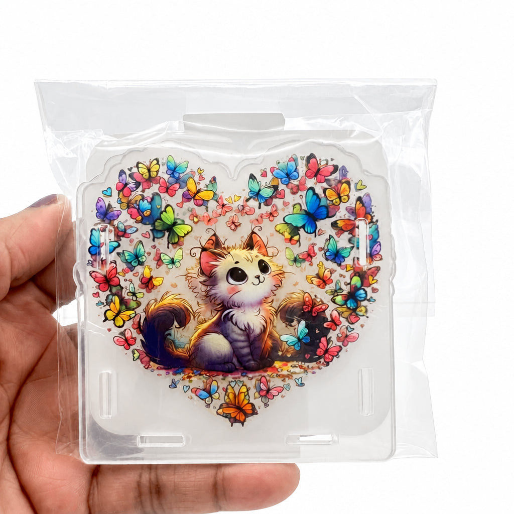 A hand holding a clear plastic bag containing a square acrylic stand or plaque featuring a cute illustrated cat surrounded by colorful butterflies on a white background.