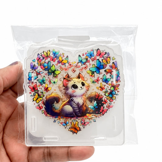 A hand holding a clear plastic bag containing a square acrylic stand or plaque featuring a cute illustrated cat surrounded by colorful butterflies on a white background.