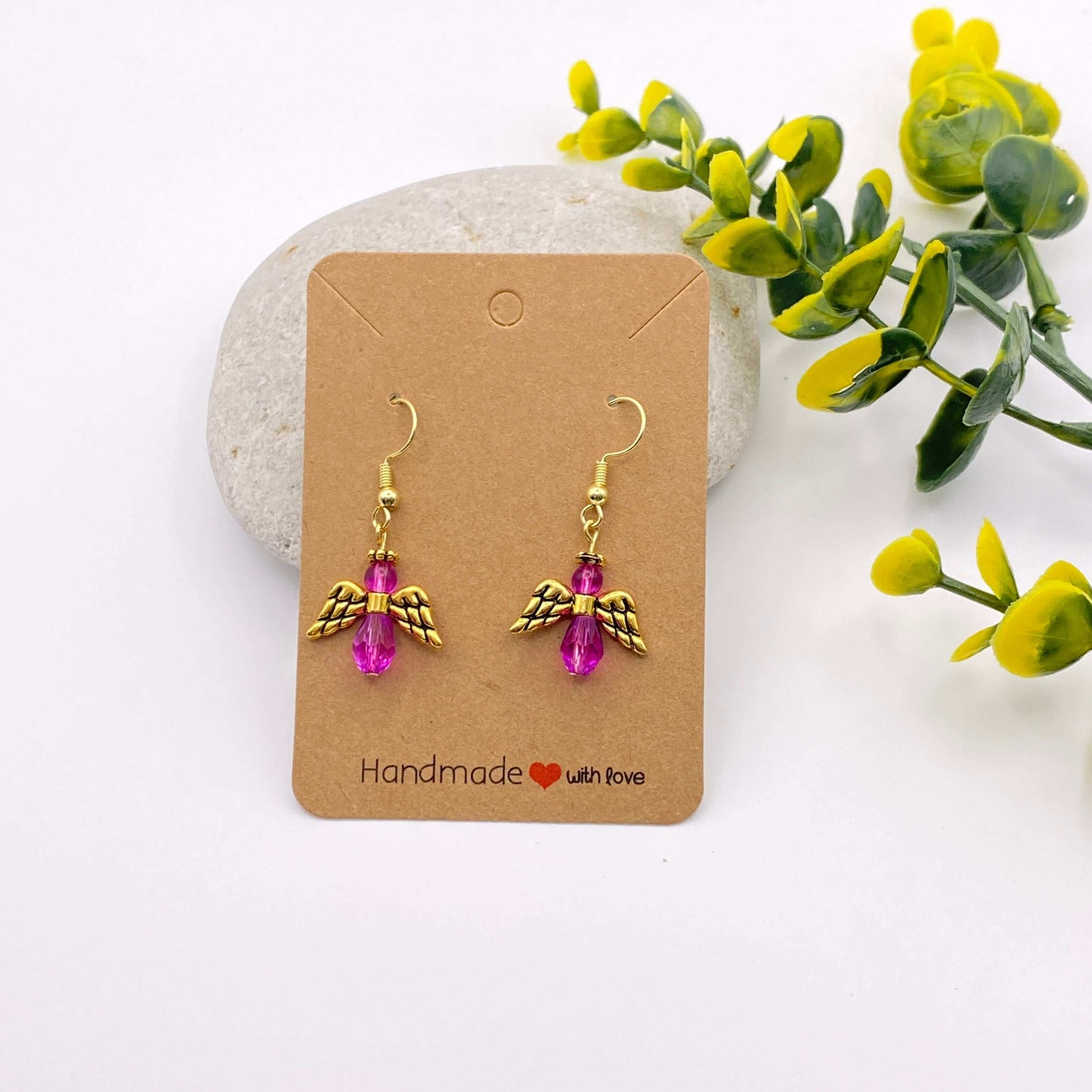 Pair of gold angel wing earrings with pink beads on a 'Handmade with Love' card, surrounded by greenery.