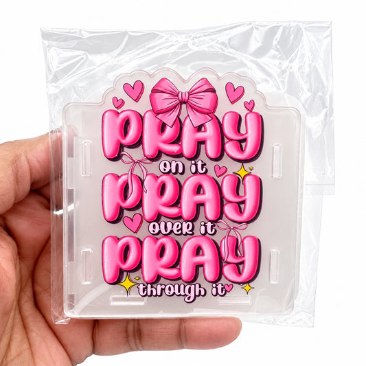 A hand holding a clear plastic case containing a pink acrylic plaque with the text 'PRAY on it PRAY over it PRAY through it' in bold pink letters, decorated with pink bows, hearts, and small yellow stars.