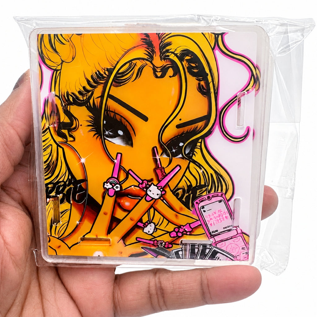 A hand holding a clear plastic-wrapped square acrylic print featuring a stylized illustrated face with orange, pink, black, and yellow tones.