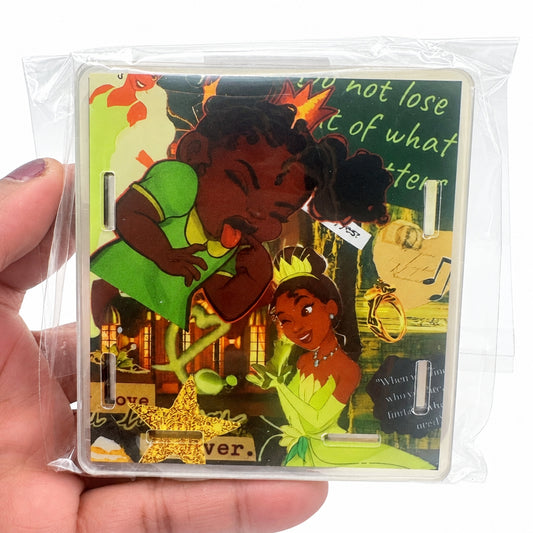 A hand holding a clear plastic-wrapped square art print featuring illustrated characters inspired by Tiana and公主, with decorative text and collage-style artwork on a white background.