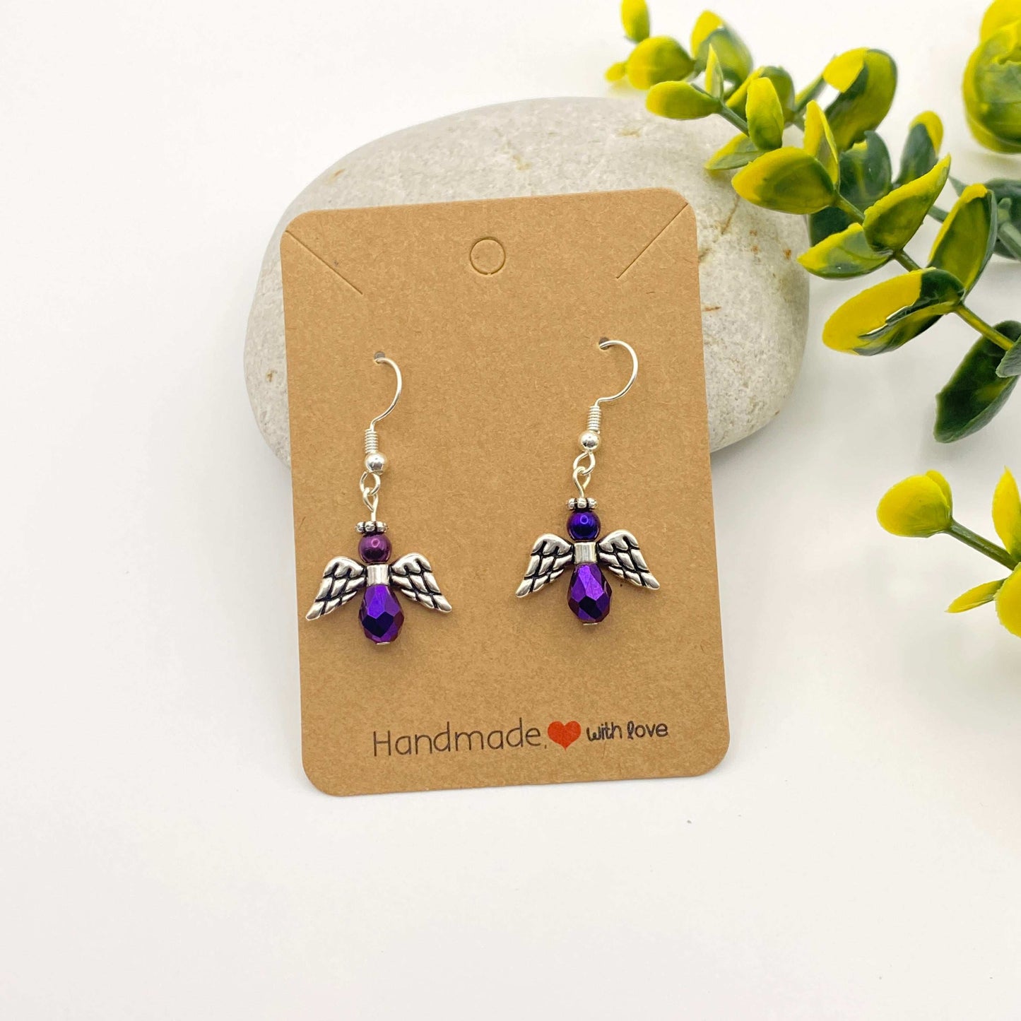 Pair of angel-winged earrings with purple beads on a 'Handmade with Love' card, with a natural stone and plant in the background.