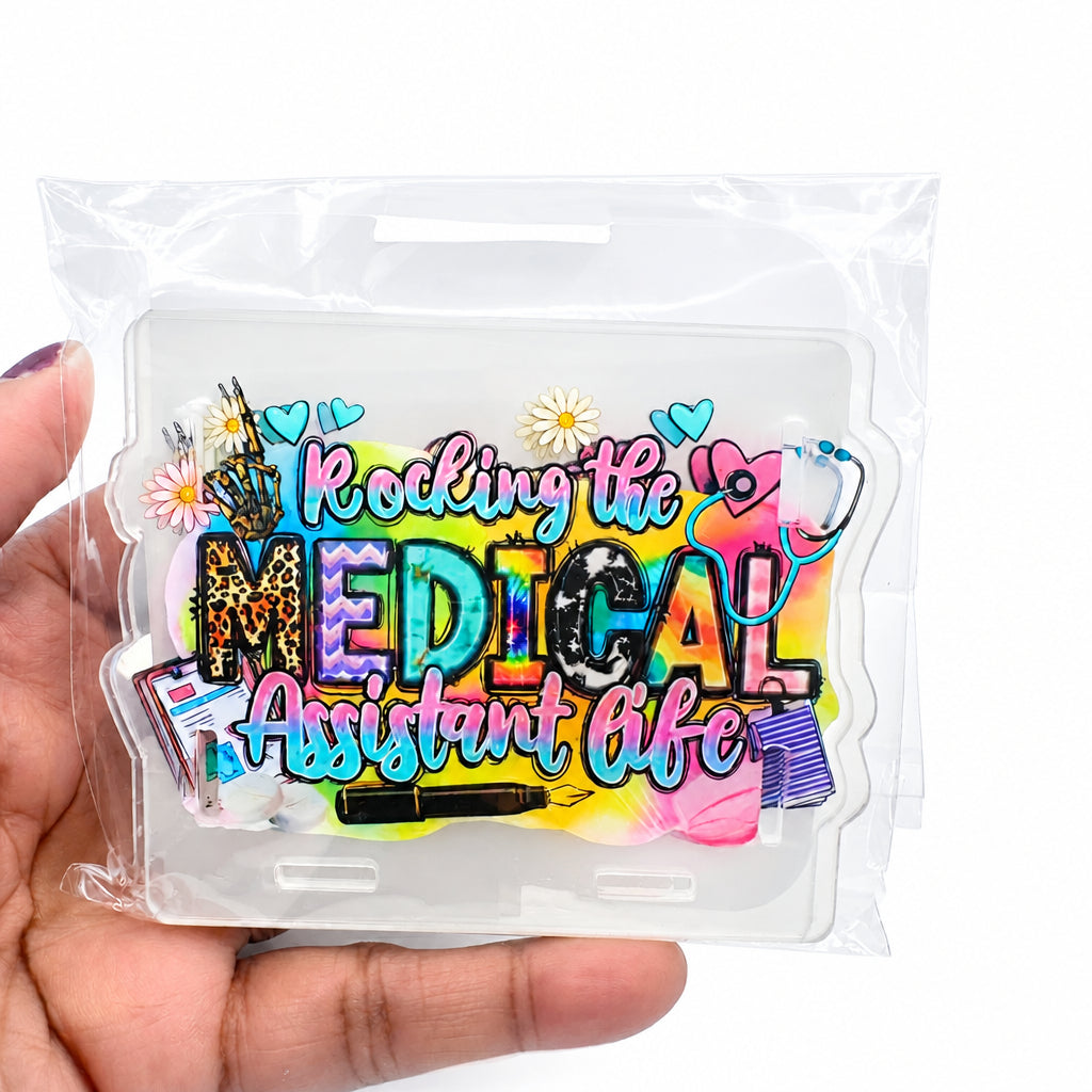 A hand holding a clear plastic bag containing a colorful acrylic plaque that reads 'Rocking the Medical Assistant Life' with medical-themed doodles and text.