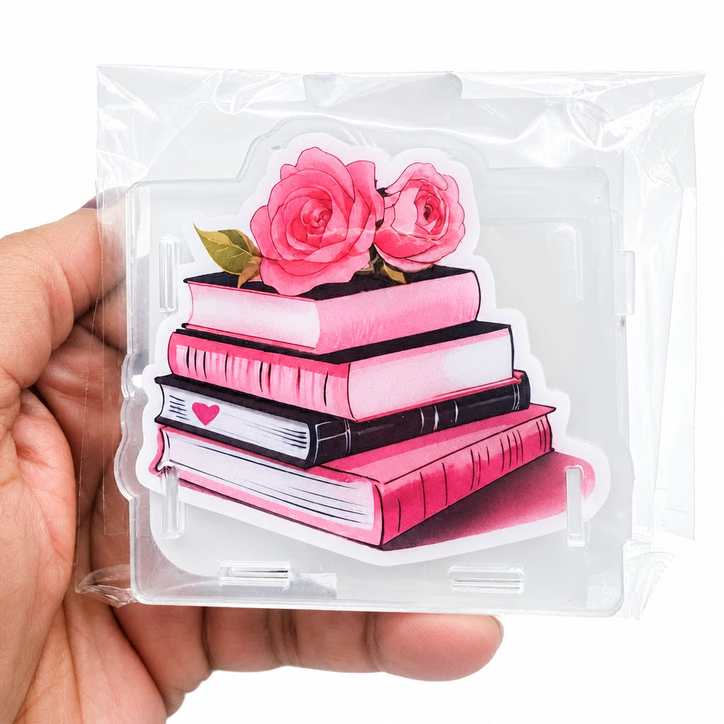 A hand holding a clear plastic sleeve containing a die-cut sticker or acrylic plaque featuring a stack of pink and black books topped with two pink roses, against a plain white background.