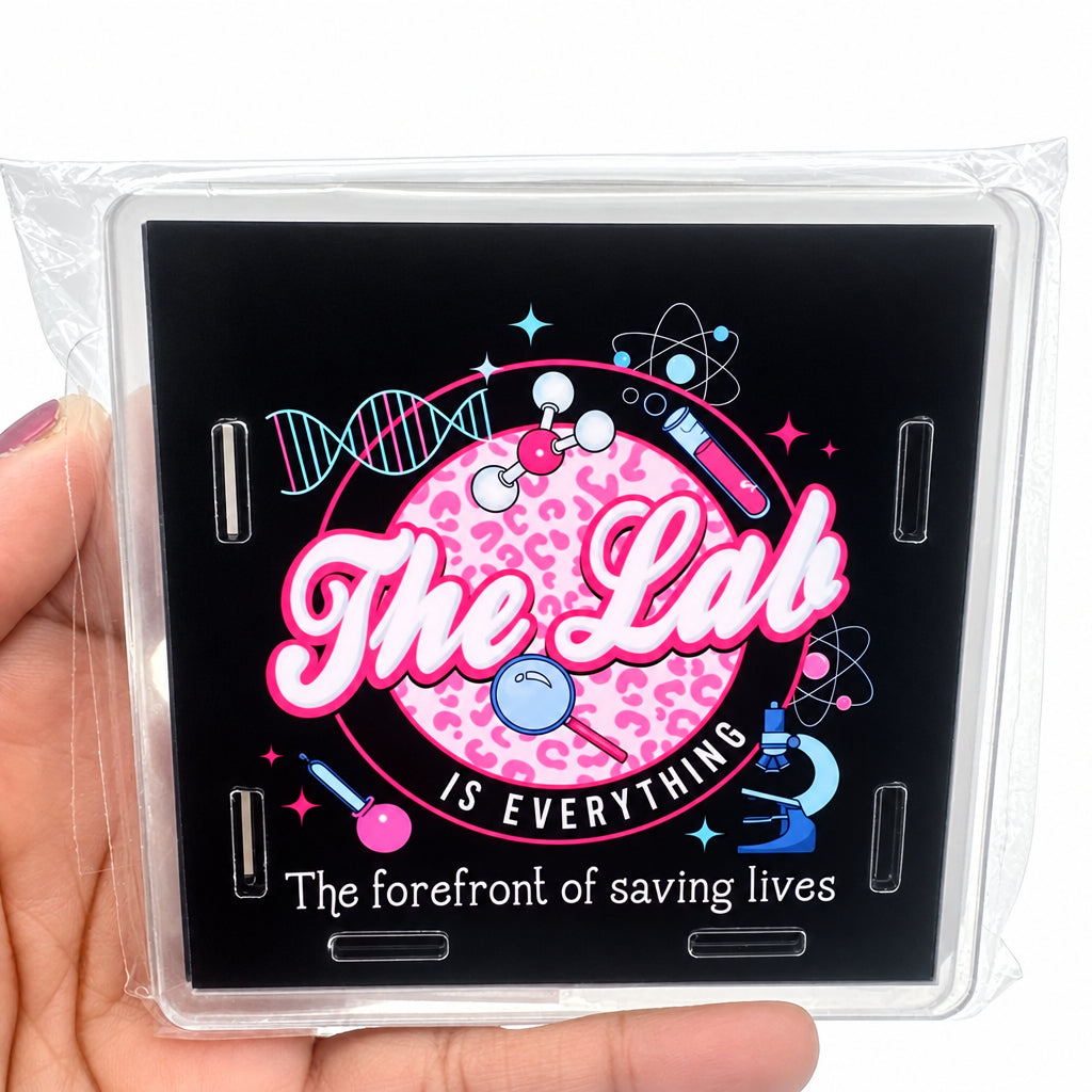 A hand holding a clear plastic-wrapped square product featuring a black background with a pink circular graphic and the words 'The Lab IS EVERYTHING' and 'The forefront of saving lives.'.