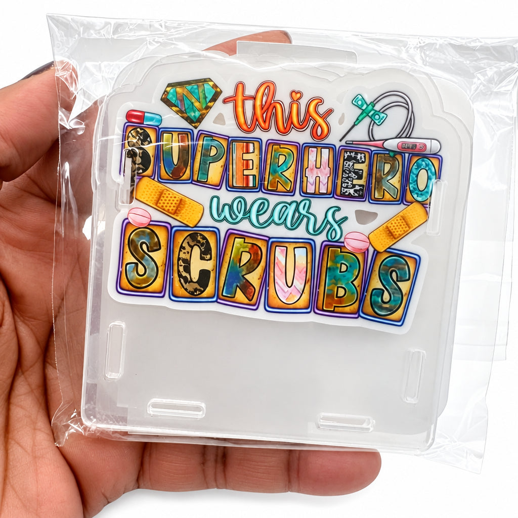 A hand holding a clear plastic case containing a die-cut sticker that reads 'this superhero wears scrubs' in colorful retro-style lettering with bandages, pencils, and medical-themed doodles.